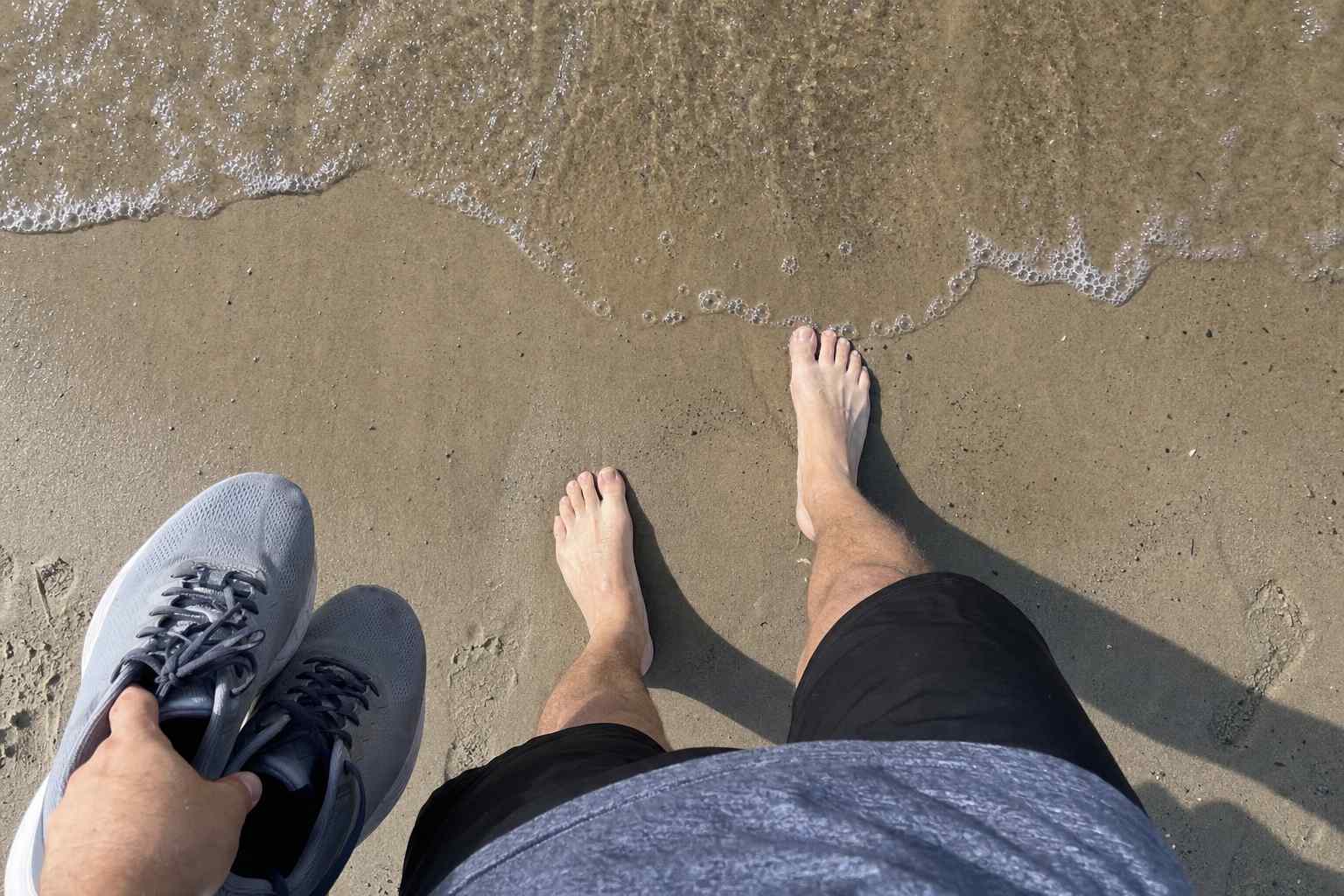 A first-person view looking down at bare feet on wet beach sand, running shoes held in hand