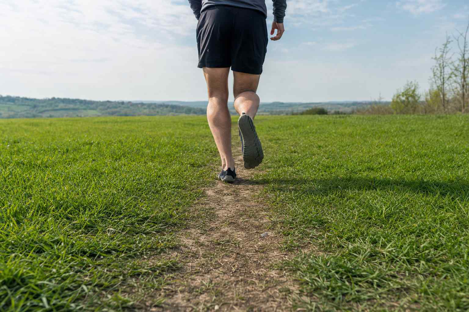 Lower legs and feet seen from behind in thin minimalist shoes, running down a path through a grass field