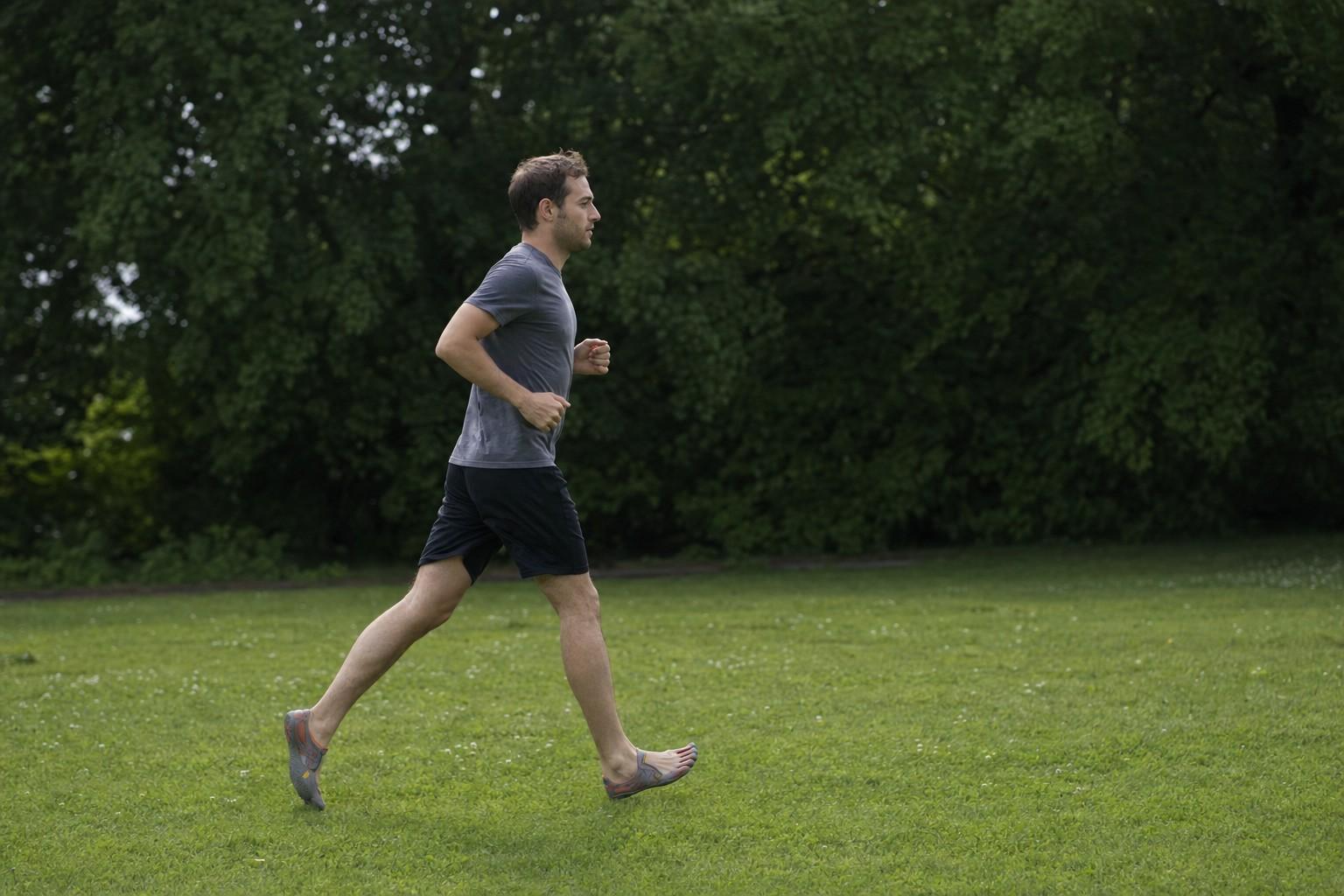 A person jogging across a grassy park in minimalist toe-style shoes, side view at mid distance